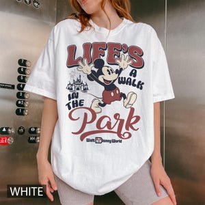K&ouml;nnte beinhalten: Wei&szlig;es T-Shirt mit Mickey Mouse-Grafik und dem Text "LIFE'S A WALK IN THE PARK" und "Walt Disney World". Das Design beinhaltet ein Schloss und Feuerwerk. Das T-Shirt hat einen Rundhalsausschnitt.