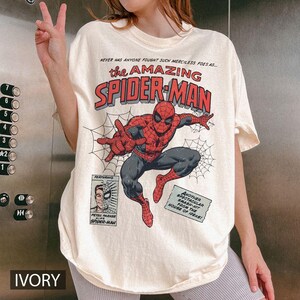 Puede incluir: Camiseta de color marfil con un gr&aacute;fico de c&oacute;mic de Spider-Man en rojo, negro y gris. El texto "the AMAZING SPIDER-MAN" se muestra en rojo y gris. El texto adicional incluye "NEVER HAS ANYONE FOUGHT SUCH MERCILESS FOES AS..." y "FEATURING PETER PARKER ALIAS SPIDER-MAN."
