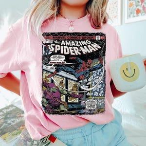May include: Pink crew neck sweatshirt featuring a vintage comic book cover of The Amazing Spider-Man. The comic book cover shows Spider-Man in action, with the Green Goblin. The sweatshirt has a relaxed fit.