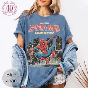 Puede incluir: Camiseta azul descolorida con un gr&aacute;fico de Spider-Man frente a un horizonte urbano. El texto "Marvel Studios Spider-Man Brand New Day" est&aacute; impreso en rojo y blanco. La camiseta es de corte holgado.