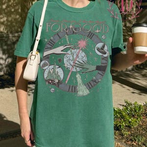 Puede incluir: Camiseta verde con un dise&ntilde;o gr&aacute;fico circular que presenta manos, una varita m&aacute;gica y un dise&ntilde;o de reloj. El texto "Because I knew you, I have been changed for good" est&aacute; impreso encima del gr&aacute;fico. Un bolso blanco es visible a la izquierda.