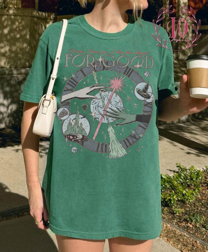 Puede incluir: Camiseta verde con un dise&ntilde;o gr&aacute;fico circular que presenta manos, una varita m&aacute;gica y un dise&ntilde;o de reloj. El texto "Because I knew you, I have been changed for good" est&aacute; impreso encima del gr&aacute;fico. Un bolso blanco es visible a la izquierda.