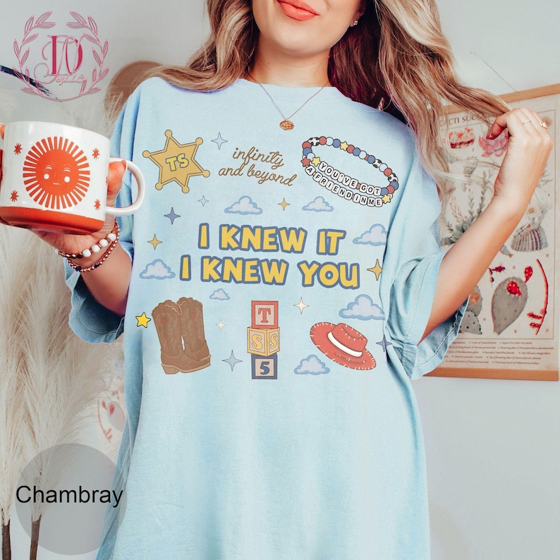 Puede incluir: Una camiseta azul claro con un estampado gr&aacute;fico con el texto I KNEW IT I KNEW YOU, infinity and beyond, y YOU'VE GOT A FRIEND IN ME. El dise&ntilde;o incluye una estrella de sheriff amarilla, una pulsera de cuentas, botas vaqueras, bloques y un sombrero.