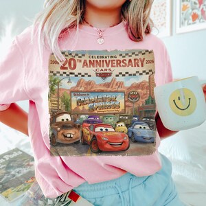 Puede incluir: Una camiseta rosa claro con un estampado gr&aacute;fico que celebra el 20 aniversario de la pel&iacute;cula Cars. El dise&ntilde;o muestra el letrero de Radiator Springs y varios personajes, incluidos Lightning McQueen y Mate, con el texto Celebrating 20th Anniversary 2006-2026.