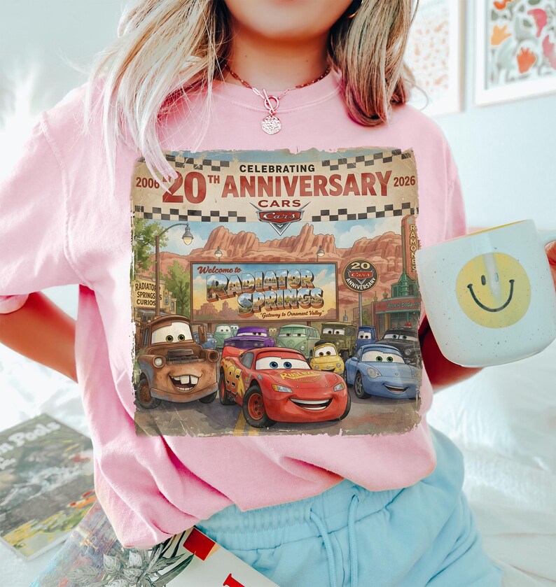 Puede incluir: Una camiseta rosa claro con un estampado gr&aacute;fico que celebra el 20 aniversario de la pel&iacute;cula Cars. El dise&ntilde;o muestra el letrero de Radiator Springs y varios personajes, incluidos Lightning McQueen y Mate, con el texto Celebrating 20th Anniversary 2006-2026.