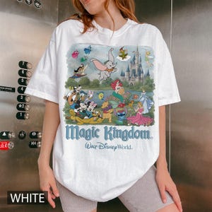 May include: A white short-sleeve t-shirt featuring a colorful graphic print of various Disney characters, a castle, and the text Magic Kingdom Walt Disney World. The design includes Dumbo, Mickey Mouse, Minnie Mouse, Ariel, and other animated figures.