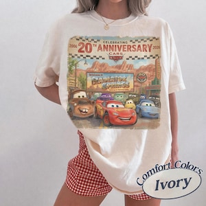 Puede incluir: Una camiseta de manga corta color marfil con un estampado gr&aacute;fico que celebra el 20 aniversario de la pel&iacute;cula Cars. El dise&ntilde;o incluye el texto Celebrating 20th Anniversary 2006-2026, Welcome to Radiator Springs e ilustraciones de personajes como Lightning McQueen y Mater.