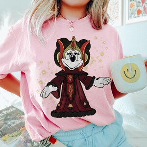 Puede incluir: Camiseta rosa claro con un personaje de dibujos animados vestido con un atuendo burdeos y dorado. El personaje lleva un tocado y una t&uacute;nica larga. En la imagen se sostiene una taza blanca con una carita sonriente amarilla.