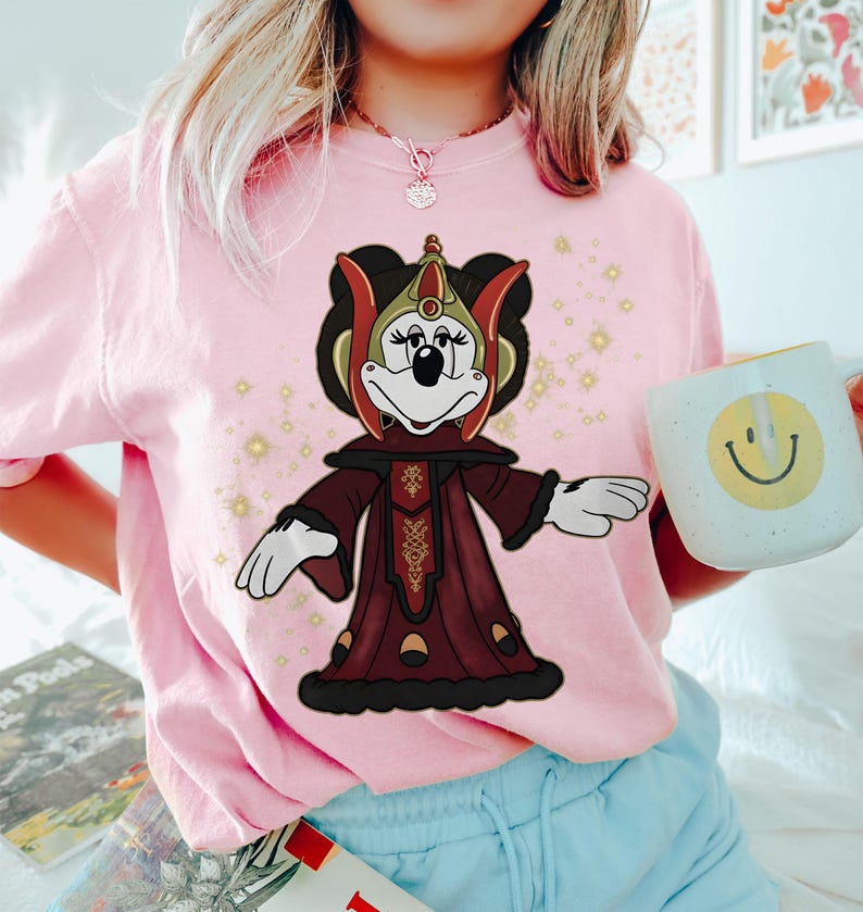 Puede incluir: Camiseta rosa claro con un personaje de dibujos animados vestido con un atuendo burdeos y dorado. El personaje lleva un tocado y una t&uacute;nica larga. En la imagen se sostiene una taza blanca con una carita sonriente amarilla.