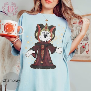 Puede incluir: Camiseta Chambray azul claro con un personaje de dibujos animados con un atuendo burdeos y dorado. El personaje tiene una cara negra y guantes blancos. Una taza blanca con un dise&ntilde;o de sol se sostiene en la mano izquierda.