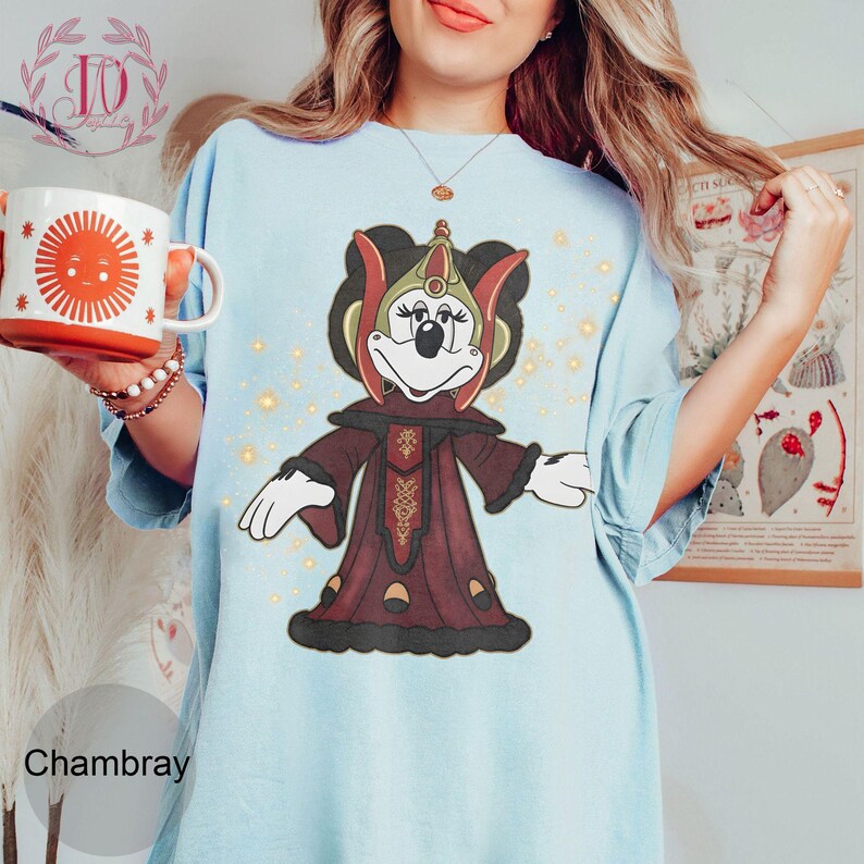 Puede incluir: Camiseta Chambray azul claro con un personaje de dibujos animados con un atuendo burdeos y dorado. El personaje tiene una cara negra y guantes blancos. Una taza blanca con un dise&ntilde;o de sol se sostiene en la mano izquierda.