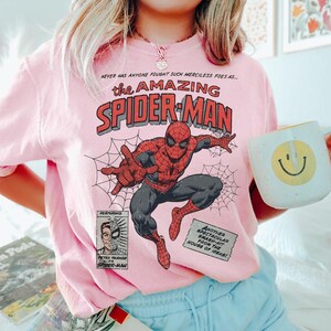 Puede incluir: Una sudadera rosa claro con un dise&ntilde;o de c&oacute;mic de Spider-Man. El dise&ntilde;o incluye las palabras "the AMAZING SPIDER-MAN" en rojo y negro, con Spider-Man en una pose din&aacute;mica. La sudadera tambi&eacute;n tiene texto adicional y una peque&ntilde;a imagen de Peter Parker.