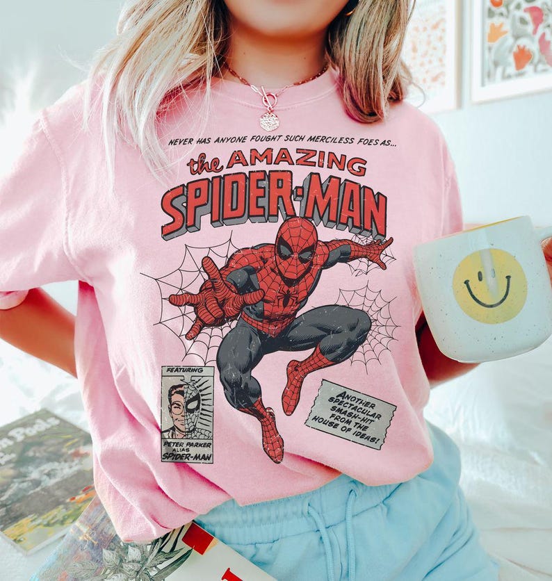 Puede incluir: Una sudadera rosa claro con un dise&ntilde;o de c&oacute;mic de Spider-Man. El dise&ntilde;o incluye las palabras "the AMAZING SPIDER-MAN" en rojo y negro, con Spider-Man en una pose din&aacute;mica. La sudadera tambi&eacute;n tiene texto adicional y una peque&ntilde;a imagen de Peter Parker.