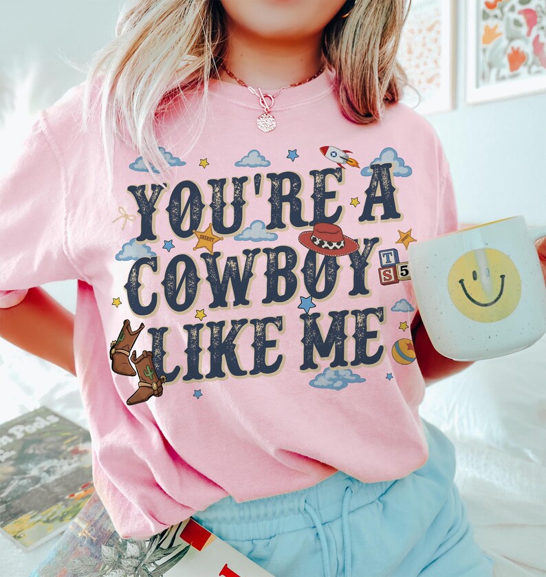 May include: A light pink short-sleeve t-shirt featuring the dark blue text "YOU'RE A COWBOY LIKE ME" surrounded by graphic illustrations of a cowboy hat, boots, clouds, stars, a rocket, and alphabet blocks with the letters T and S.