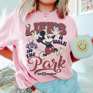 K&ouml;nnte beinhalten: Hellrosa T-Shirt mit Mickey Mouse und dem Text "LIFE'S A WALK IN THE PARK" in Braun und Wei&szlig;. Das Walt Disney World Logo ist ebenfalls zu sehen. Eine wei&szlig;e Tasse mit einem gelben Smiley wird im Bild gehalten.