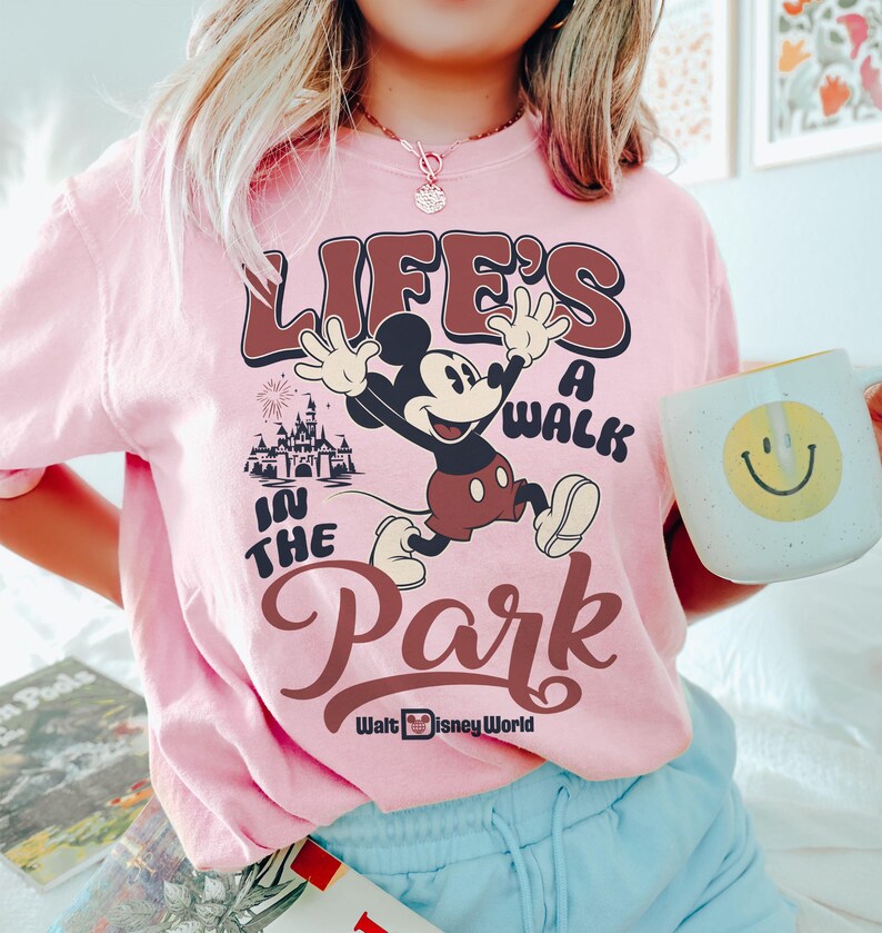 K&ouml;nnte beinhalten: Hellrosa T-Shirt mit Mickey Mouse und dem Text "LIFE'S A WALK IN THE PARK" in Braun und Wei&szlig;. Das Walt Disney World Logo ist ebenfalls zu sehen. Eine wei&szlig;e Tasse mit einem gelben Smiley wird im Bild gehalten.