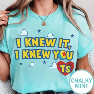 Op de afbeelding: Een licht mintgroen t-shirt met de gele tekst I KNEW IT, I KNEW YOU, een kleurrijke balafbeelding die de letter O vervangt, een rood hart met de gele letters TS, en kleine witte wolken en blauwe sterren op de voorzijde gedrukt.