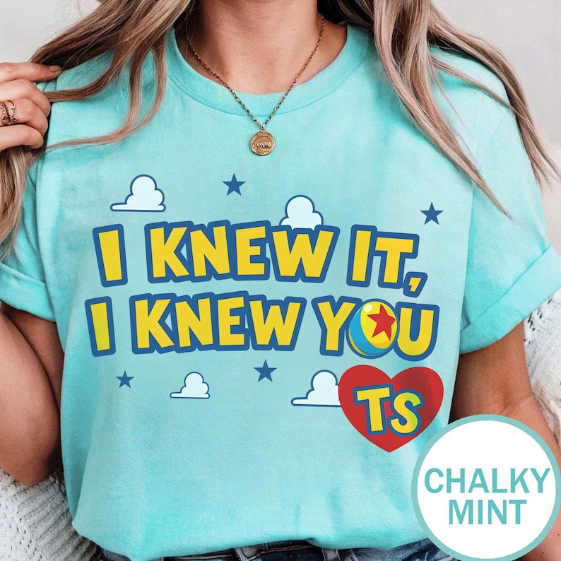 May include: A light mint green t-shirt featuring the yellow text I KNEW IT, I KNEW YOU with a colourful ball graphic replacing the letter O, a red heart containing the yellow letters TS, and small white clouds and blue stars printed on the front.