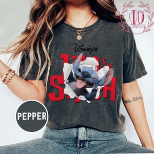 Op de afbeelding: Donkergrijs T-shirt met een afbeelding van Disney's Stitch die door een gescheurd papiereffect breekt. De tekst "Disney's Stitch" is rood. Een casual kledingstuk.