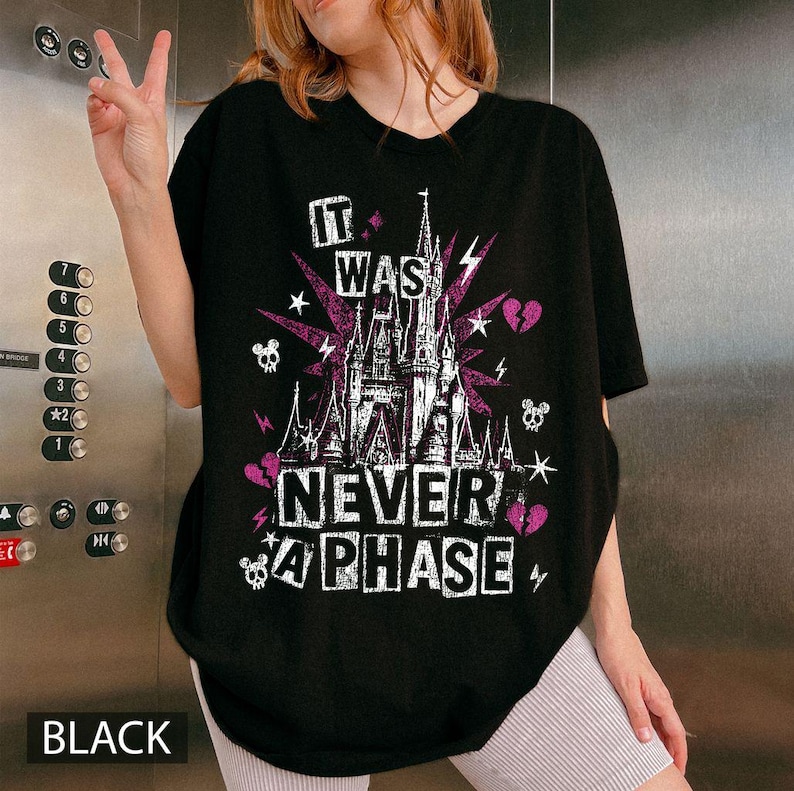 May include: Black t-shirt with a graphic design featuring a castle, lightning bolts, skulls, and the text "IT WAS NEVER A PHASE". The design incorporates purple and white elements. The word "BLACK" is at the bottom.