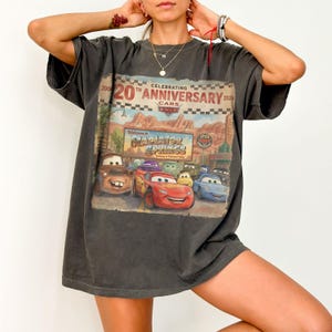 Puede incluir: Una camiseta gris carb&oacute;n de manga corta con un estampado gr&aacute;fico que celebra el 20 aniversario de la pel&iacute;cula Cars. El dise&ntilde;o incluye el texto Celebrating 20th Anniversary Cars 2006-2026 y Welcome to Radiator Springs con varios personajes.