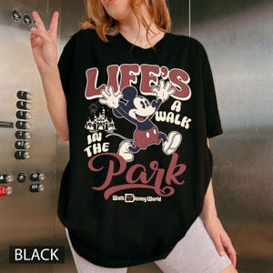 K&ouml;nnte beinhalten: Schwarzes T-Shirt mit einer Mickey Mouse-Grafik und dem Text "LIFE'S A WALK IN THE PARK" und "Walt Disney World." Das Design beinhaltet ein Schloss und Feuerwerk. Das Wort "BLACK" ist unten auf dem T-Shirt aufgedruckt.