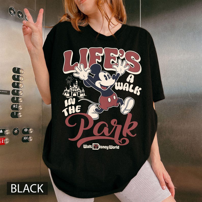 K&ouml;nnte beinhalten: Schwarzes T-Shirt mit einer Mickey Mouse-Grafik und dem Text "LIFE'S A WALK IN THE PARK" und "Walt Disney World." Das Design beinhaltet ein Schloss und Feuerwerk. Das Wort "BLACK" ist unten auf dem T-Shirt aufgedruckt.