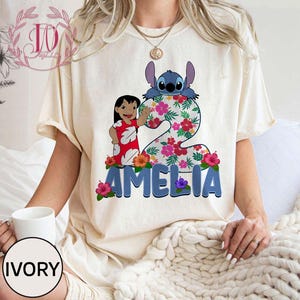 Op de afbeelding: Ivoor kleurig T-shirt met een kleurrijke afbeelding van Lilo en Stitch. Het cijfer 2 is versierd met bloemen en de naam "AMELIA" staat eronder. Het shirt heeft korte mouwen en een relaxte pasvorm.