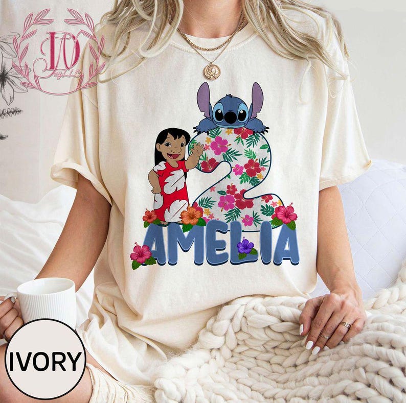 Op de afbeelding: Ivoor kleurig T-shirt met een kleurrijke afbeelding van Lilo en Stitch. Het cijfer 2 is versierd met bloemen en de naam "AMELIA" staat eronder. Het shirt heeft korte mouwen en een relaxte pasvorm.