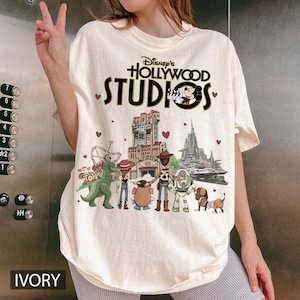 Pode incluir: Camiseta cor marfim com o texto "Disney's Hollywood Studios" e Mickey Mouse. A camisa apresenta v&aacute;rios personagens da Disney e atra&ccedil;&otilde;es do parque. A palavra "IVORY" est&aacute; impressa na parte inferior.
