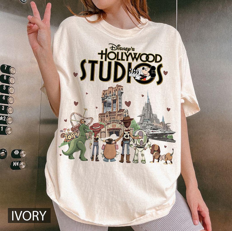 Pode incluir: Camiseta cor marfim com o texto "Disney's Hollywood Studios" e Mickey Mouse. A camisa apresenta v&aacute;rios personagens da Disney e atra&ccedil;&otilde;es do parque. A palavra "IVORY" est&aacute; impressa na parte inferior.