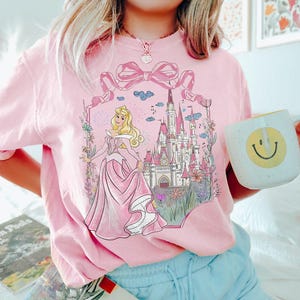 May include: Pink sweatshirt featuring a graphic of a princess in a pink gown, a castle, and floral accents. The design is framed by a large pink bow. A white mug with a yellow smiley face is held in the hand.