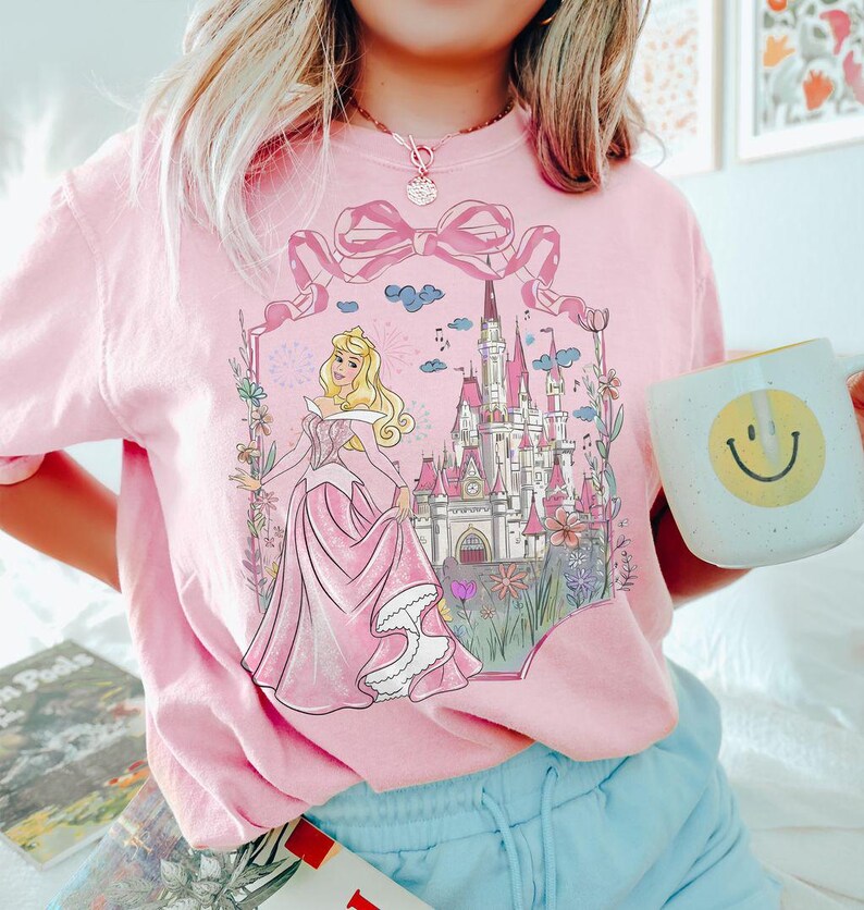 May include: Pink sweatshirt featuring a graphic of a princess in a pink gown, a castle, and floral accents. The design is framed by a large pink bow. A white mug with a yellow smiley face is held in the hand.