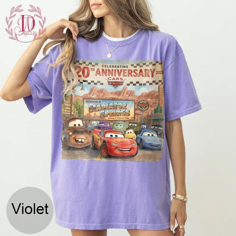 Puede incluir: Una camiseta violeta de manga corta con un estampado gr&aacute;fico que celebra el 20 aniversario de la pel&iacute;cula Cars. El dise&ntilde;o muestra el letrero de bienvenida de Radiator Springs, Rayo McQueen, Mate y otros personajes con un fondo des&eacute;rtico.