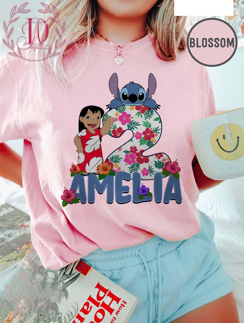 Op de afbeelding: Roze T-shirt met een afbeelding van Lilo en Stitch, het nummer 2, bloemmotieven en de naam "AMELIA". Het ontwerp bevat Stitch en Lilo in een rode jurk. Het shirt wordt gedragen met een blauwe short.