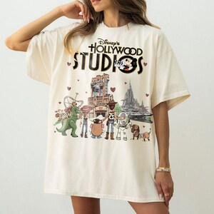 Pode incluir: Camiseta creme com o texto "Disney's Hollywood Studios", Mickey Mouse e um gr&aacute;fico de rolo de filme. A camisa apresenta um gr&aacute;fico colorido de personagens do Toy Story e pontos de refer&ecirc;ncia do parque.