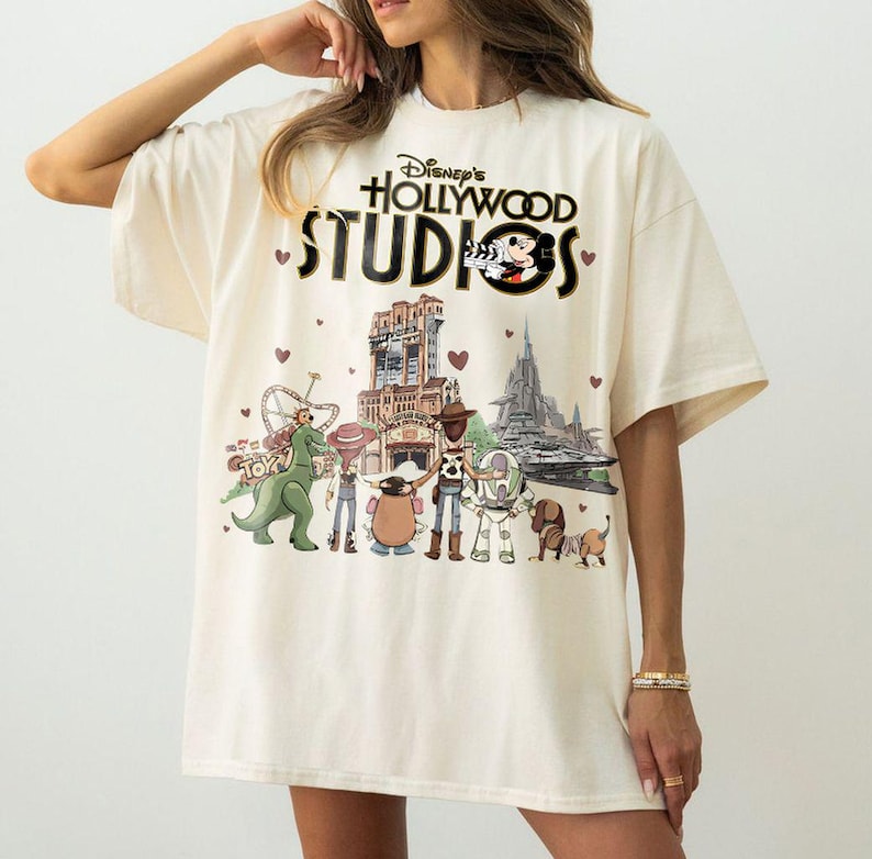 Pode incluir: Camiseta creme com o texto "Disney's Hollywood Studios", Mickey Mouse e um gr&aacute;fico de rolo de filme. A camisa apresenta um gr&aacute;fico colorido de personagens do Toy Story e pontos de refer&ecirc;ncia do parque.