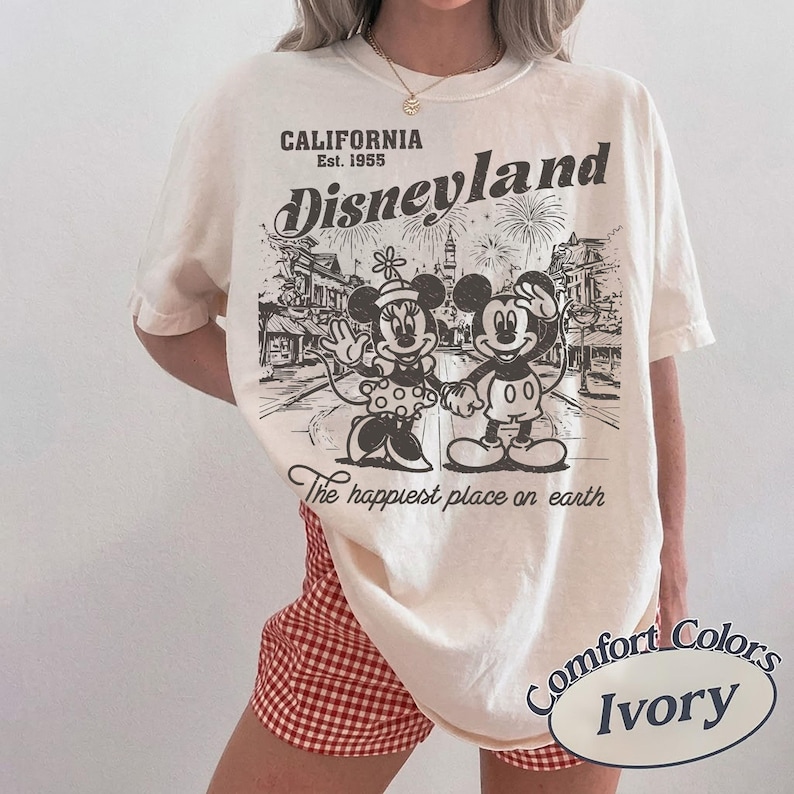 May include: Ivory-colored t-shirt featuring a vintage Disneyland graphic. The design includes Mickey and Minnie Mouse, fireworks, and the text "California Est. 1955." The phrase "The happiest place on earth" is also printed on the shirt.