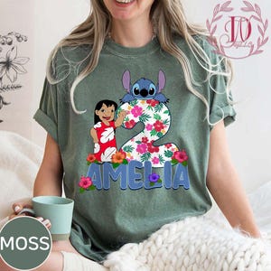 Op de afbeelding: Groen t-shirt met een kleurrijke afbeelding van Lilo en Stitch, het nummer 2, bloemendetails en de naam "AMELIA" in blauw. Het ontwerp bevat een cartoon van Lilo die zwaait en Stitch die over het nummer kijkt.