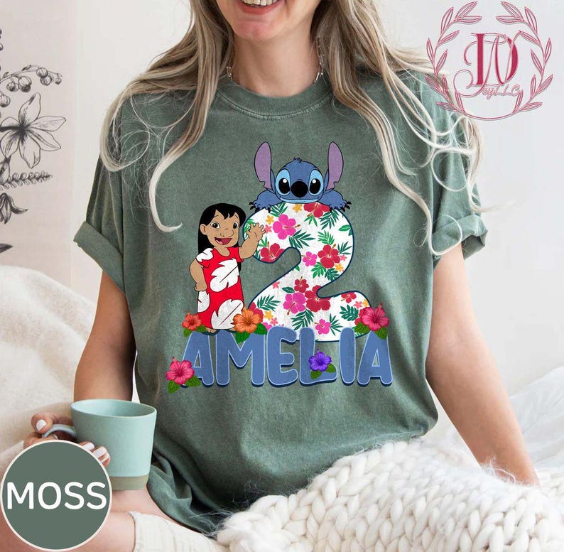 Op de afbeelding: Groen t-shirt met een kleurrijke afbeelding van Lilo en Stitch, het nummer 2, bloemendetails en de naam "AMELIA" in blauw. Het ontwerp bevat een cartoon van Lilo die zwaait en Stitch die over het nummer kijkt.