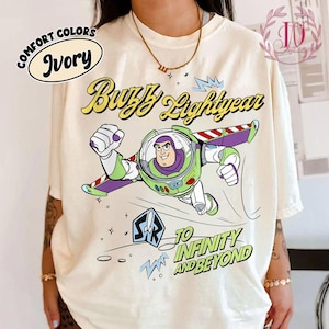 Puede incluir: Camiseta de color marfil con un gráfico de Buzz Lightyear en vuelo, con el texto "Buzz Lightyear" en amarillo y "To Infinity and Beyond" en azul. La camiseta también tiene las palabras "Comfort Colors" e "Ivory" en un diseño circular.