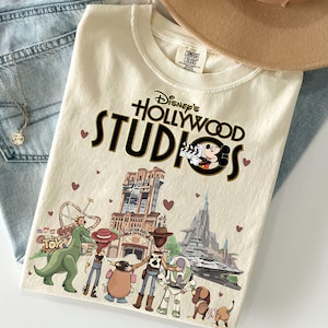 Pode incluir: Camiseta creme do Disney's Hollywood Studios com um gr&aacute;fico com Mickey Mouse, o logotipo do parque e v&aacute;rios personagens da Disney. A camisa est&aacute; dobrada sobre jeans azuis e um chap&eacute;u bege.