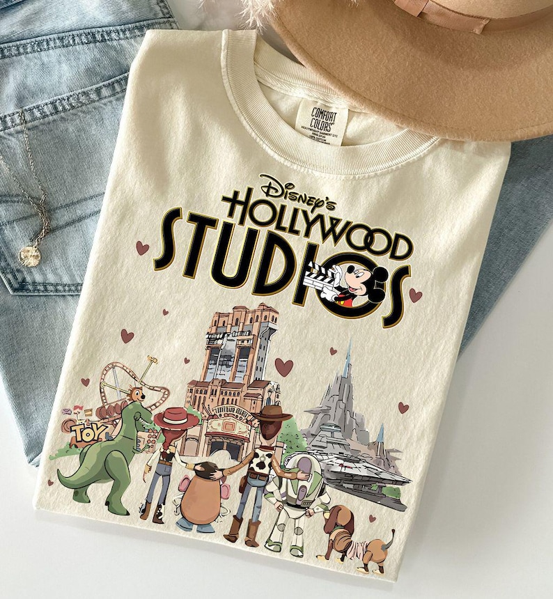 Pode incluir: Camiseta creme do Disney's Hollywood Studios com um gr&aacute;fico com Mickey Mouse, o logotipo do parque e v&aacute;rios personagens da Disney. A camisa est&aacute; dobrada sobre jeans azuis e um chap&eacute;u bege.