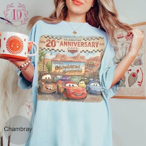 Puede incluir: Una camiseta azul claro con un estampado gr&aacute;fico que celebra el 20 aniversario de la pel&iacute;cula Cars. El dise&ntilde;o muestra el texto Celebrating 20th Anniversary 2006-2026, Radiator Springs y varios personajes, incluido Rayo McQueen.