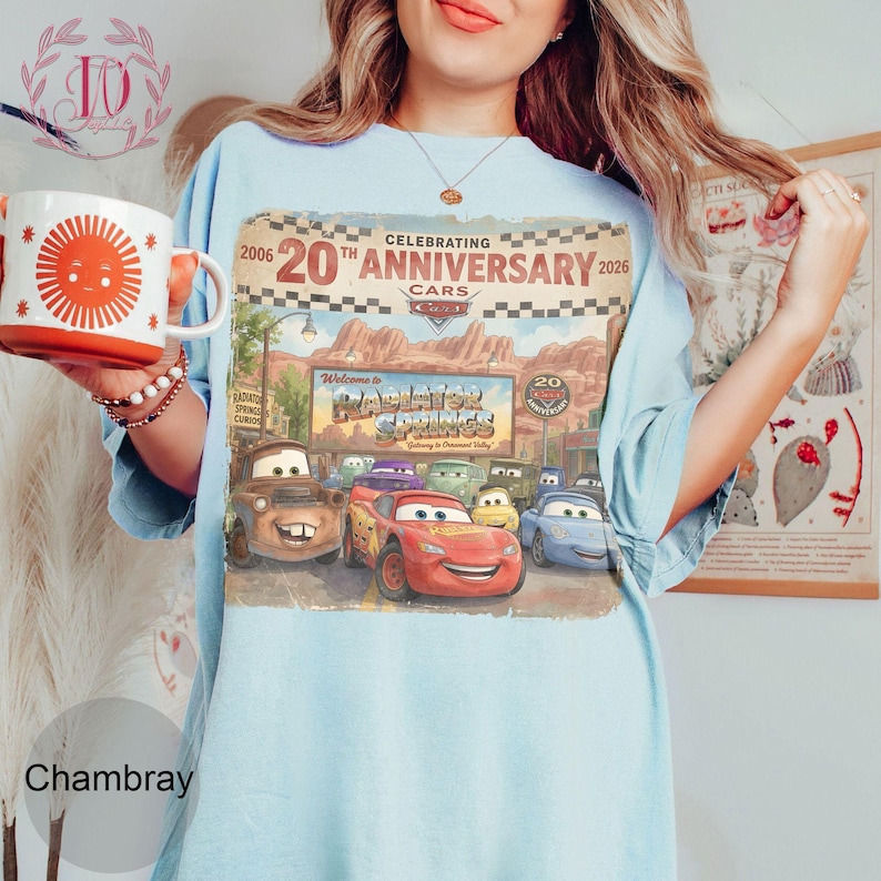 Puede incluir: Una camiseta azul claro con un estampado gr&aacute;fico que celebra el 20 aniversario de la pel&iacute;cula Cars. El dise&ntilde;o muestra el texto Celebrating 20th Anniversary 2006-2026, Radiator Springs y varios personajes, incluido Rayo McQueen.
