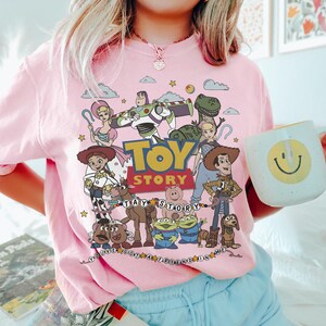 May include: A light pink t-shirt featuring a colorful graphic of Toy Story characters including Woody, Buzz Lightyear, Jessie, and Rex. The design includes the Toy Story logo, the text You've Got A Friend In Me, and various stars and clouds.