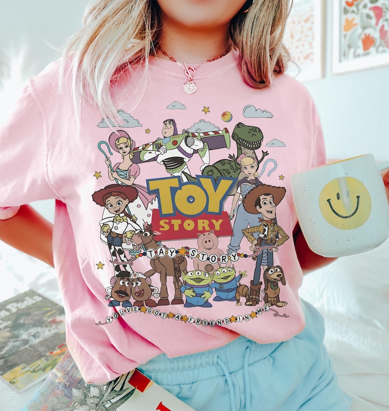 May include: A light pink t-shirt featuring a colorful graphic of Toy Story characters including Woody, Buzz Lightyear, Jessie, and Rex. The design includes the Toy Story logo, the text You've Got A Friend In Me, and various stars and clouds.
