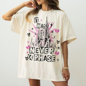 May include: Cream-colored oversized t-shirt with a graphic design featuring a castle, lightning bolts, stars, and the text "IT WAS NEVER A PHASE" in black and pink. The design has a glittery effect.