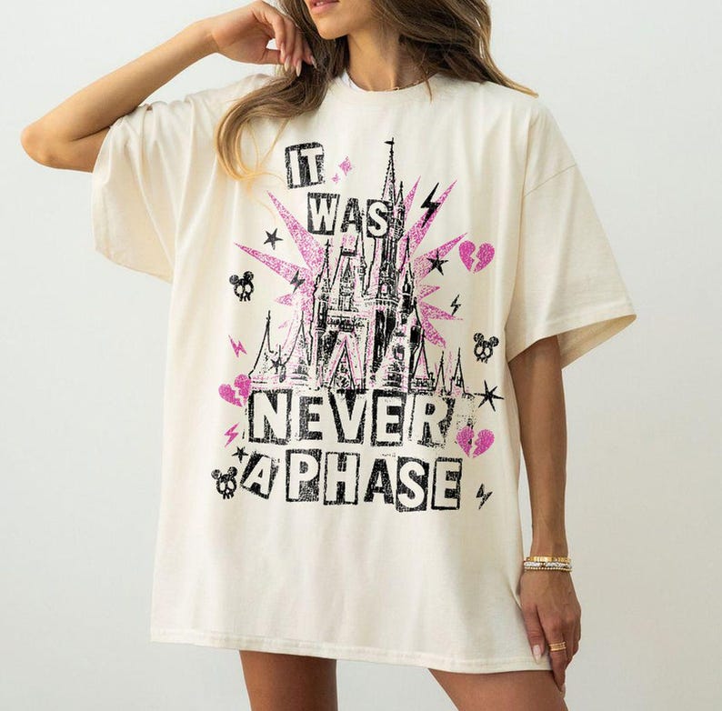 May include: Cream-colored oversized t-shirt with a graphic design featuring a castle, lightning bolts, stars, and the text "IT WAS NEVER A PHASE" in black and pink. The design has a glittery effect.