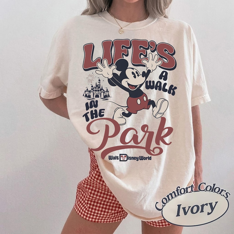 K&ouml;nnte beinhalten: Elfenbeinfarbenes T-Shirt mit einer Mickey Mouse-Grafik und dem Text "Life's a Walk in the Park" in Rot und Blau. Das Design beinhaltet ein Schloss und die Aufschrift "Walt Disney World". Das Shirt geh&ouml;rt zur Comfort Colors-Kollektion.